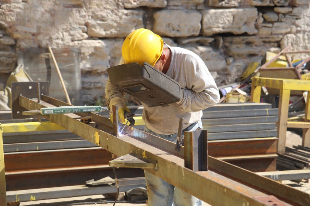 Welder wearing safety gear works with metal beams outdoors on a construction site.